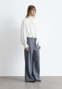 Weißes Langarmbluse mit Schleifendetail, kombiniert mit grauen, weit geschnittenen Hosen aus strukturiertem Stoff, ergänzt durch einen weißen Gürtel und schwarze Schuhe.