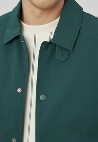Veste vert foncé avec un col pointu, des boutons-pression et un tissu texturé. Portée sur une chemise claire à rayures verticales.