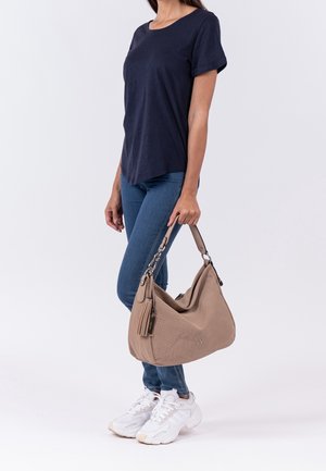 Sac à bandoulière texturé couleur beige avec une forme courbée, une ferrure argentée et un détail pompon, assorti à un t-shirt bleu marine foncé et un jean bleu.