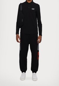 Černé dlouhé polo s logem EA7, kombinované s černými tepláky se šedými bočními panely a červenými textovými akcenty. Bílí sneakers dotvářejí celý outfit.