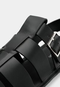 Μαύρο δερμάτινο σανδάλι με φαρδιές διασταυρούμενες λουρίδες και ασημένια μεταλλική αγκράφα για ρύθμιση. Λεία υφή με μινιμαλιστικές σχεδιαστικές λεπτομέρειες.