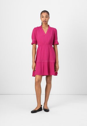 Fuchsia kurzes Kleid mit Puffärmeln, V-Ausschnitt und besticktem TaillenDetail. Das Modell trägt schwarze Ballerinas. Minimale Hintergrundgestaltung.