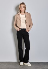 Veste courte tan avec des boutons double-breasted, t-shirt graphique crème, pantalons noirs et baskets blanches. Tissu lisse, coupe décontractée.
