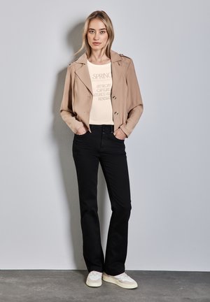 Tan cropped jakke med dobbeltknaplukning, cremet grafisk t-shirt, sorte bukser og hvide sneakers. Glat stof, afslappet pasform.