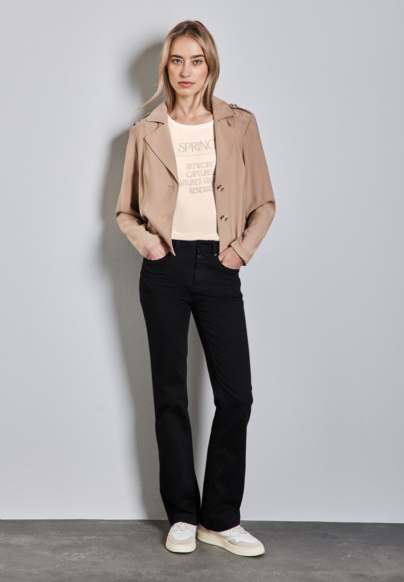 Tan cropped jacka med dubbelknäppta knappar, krämfärgad t-shirt med grafiskt tryck, svarta byxor och vita sneakers. Slät tygkvalitet, avslappnad passform.
