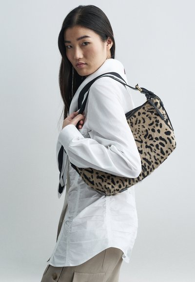 Kobieta w białej, marszczonej koszuli i beżowych spodniach, trzymająca torebkę na ramię w panterkę z czarnymi paskami na tle jednolitym.