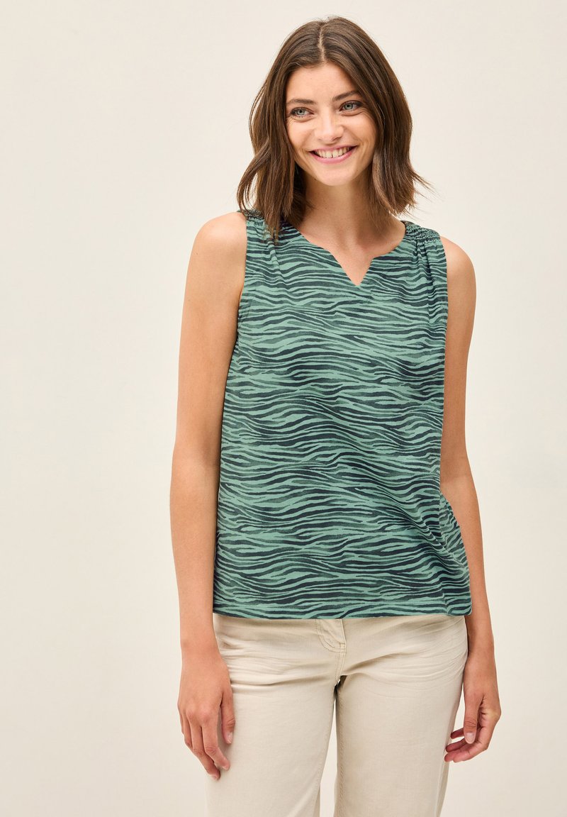 Canotta verde con un sottile motivo a strisce zebra, design a V e dettagli arricciati sulle spalle. Abbinata a pantaloni beige chiaro.