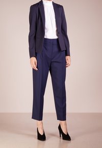 Blazer sartoriale blu navy e pantaloni coordinati in tessuto liscio. Design aderente con silhouette strutturata e scarpe nere con tacco alto.