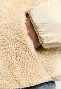 Beige fleecejacka med en texturerad, fluffig utsida och släta, enfärgade ärmar med en synlig ficköppning.