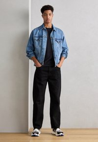 Τζάκετ από τζιν σε ανοιχτό μπλε χρώμα με δύο τσέπες στο στήθος, φορεμένο πάνω από μαύρο πουκάμισο, συνδυασμένο με μαύρο παντελόνι και αθλητικά παπούτσια σε μαύρο και άσπρο χρώμα. Απλή φόντο.