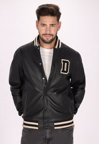 Veste varsity en cuir noir avec une fermeture par pression, col et poignets côtelés rayés, et une lettre 'D' bien visible sur la poitrine.