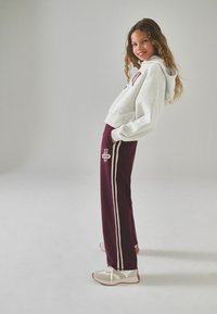 Giovane donna inclinata all'indietro, con indosso un hoodie grigio chiaro, pantaloni da jogging bordeaux con strisce bianche e sneakers beige, in piedi su uno sfondo neutro.