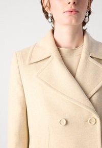 Bežový vlněný dvouřadý kabát s širokými klopami a dvěma předními kapsami. Hladká textura se jemnými skvrnami, nošený přes neutrální top.