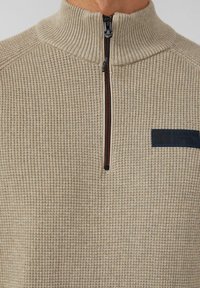 Beige Zip-Hoodie mit geripptem Muster, hohem Kragen und einem marineblauen Logopatch auf der linken Brust. Verfügt über kontrastierende Reißverschlussdetails.