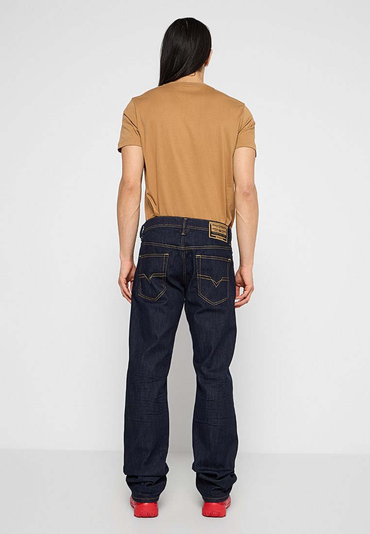 Dunkelblaue Jeans aus Denim mit gelben Nähten und einem Rückenklebeetikett, kombiniert mit einem kurzärmeligen braunen Hemd. Rückansicht, rote Schuhe sichtbar.