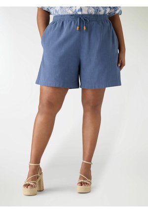 Pantaloncini di lino blu con vita elastica e coulisse indossati con sandali beige con plateau da una persona in piedi.