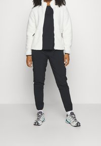 Vit fleecejacka med hög krage, i kombination med en svart skjorta och svarta smala byxor. Skorna är grå och teal färgade sportskor.