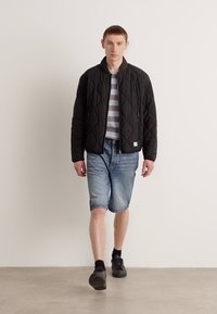 Svart quiltet bomberjakke med glidelås, stripete grå og hvit t-skjorte, denimshorts og svarte sokker med sneakers. Enkelt bakgrunn.