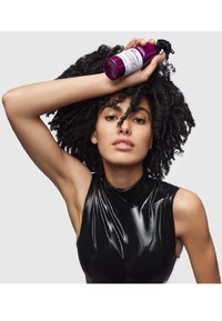 Paarse haarproductfles boven een krullend zwart haar, tegen een lichtgrijze achtergrond. Het model draagt een glanzend zwart mouwloos topje.