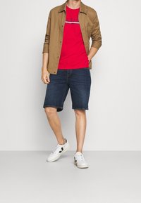 Hombre que lleva una camiseta de logo rojo, una camisa de botones beige, pantalones cortos de mezclilla oscuros y zapatillas blancas, de pie contra un fondo plano.