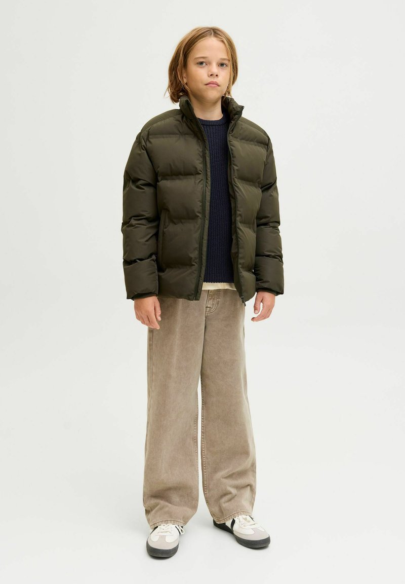 Olivengrüne Pufferjacke mit hohem Kragen, getragen über einem marineblauen Strickpullover. Kombiniert mit beigen weit geschnittenen Hosen und schwarzen Sneakers.