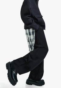 Giacca nera corta sopra pantaloni neri a gamba larga con pannello in plaid. Scarpe nere con texture e suola spessa. Design minimalista e contrasto di colori.