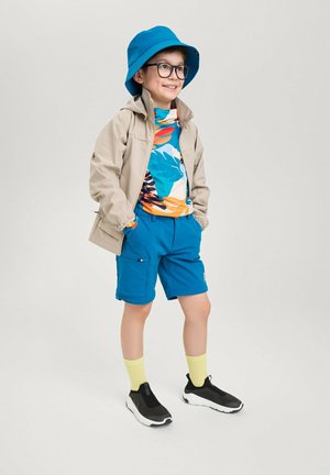 Helle beige Jacke, leuchtend blauer Bucket Hat, bunte kurzärmelige Hemd mit tropischem Muster, blaue Shorts und schwarze Schuhe mit weißen Sohlen.