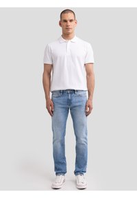 Egy férfi áll, fehér galléros pólót, világoskék farmert és fehér sportcipőt visel, egy egyszerű szürke háttér előtt.