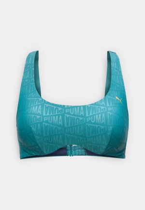 Τιρκουάζ αθλητικό σουτιέν με φαρδιές τιράντες και επαναλαμβανόμενα λογότυπα "PUMA", που διαθέτει ένα μικρό χρυσό λογότυπο Puma γάτας στη δεξιά πλευρά.