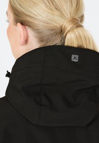 Musta takki korkealla kauluksella ja pienellä logolla takana. Materiaali näyttää sileältä, ja siinä on siistit saumat sekä jämäkkä muotoilu.