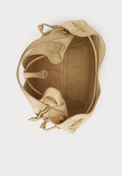 Bež semišasta torbica s prostorno notranjostjo in mehko teksturo. Ima zaobljene robove, usnjene trakove in dekorativne detajle šivov.