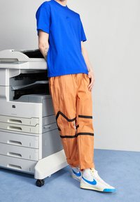 Královsky modré bavlněné tričko s malým logem, spárované s oranžovými zmačkanými kalhotami s černými geometrickými akcenty a bílými teniskami s modrými detaily.