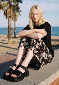 Ung kvinde med langt blondt hår sidder på en betonkant nær en strand, iført en sort skjorte, blomsterprint nederdel og sorte sandaler.