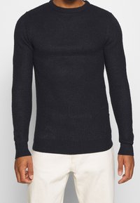 Svart stickad tröja med långa ärmar och rund halsringning, med en texturerad mönstring och ribbade muddar och nederkant.