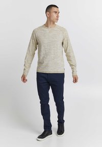 Világos bézs hosszú ujjú pulóver, texturált mintázattal, kerek nyakkivágással, és ellentétes sötétkék slim-fit nadrággal. Fekete magas szárú tornacipők.