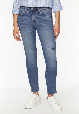 Chude jeansy w średnio niebieskim denimu z przetarciami na kolanach. Zawierają zapięcie na guzik oraz pięć kieszeni, połączone z białymi trampkami.