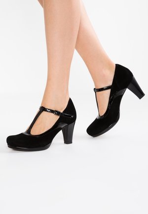 Beine, die schwarze T-Riemchen-Pumps mit abgerundeten Zehen, glänzender Schnalle und mittelhohen klobigen Absätzen vor weißem Hintergrund tragen.