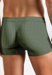 Man in een strak passend olijfgroen boxershort met een klein vierkant patroon, van achteren gezien tegen een witte achtergrond.