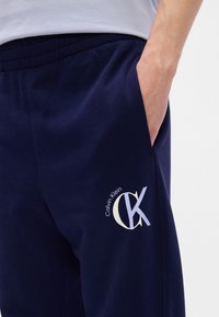 Tengerészkék melegítőnadrág puha anyagból, rugalmas derekú és oldalsó zsebes kivitelben. Az alsó lábszáron egy fehér Calvin Klein logó látható.