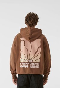 Barna kapucnis pulóver, amelyen egy nagy, stilizált pillangó grafika látható rózsaszín és krém színekben, alatta a "LOST YOUTH" felirattal.