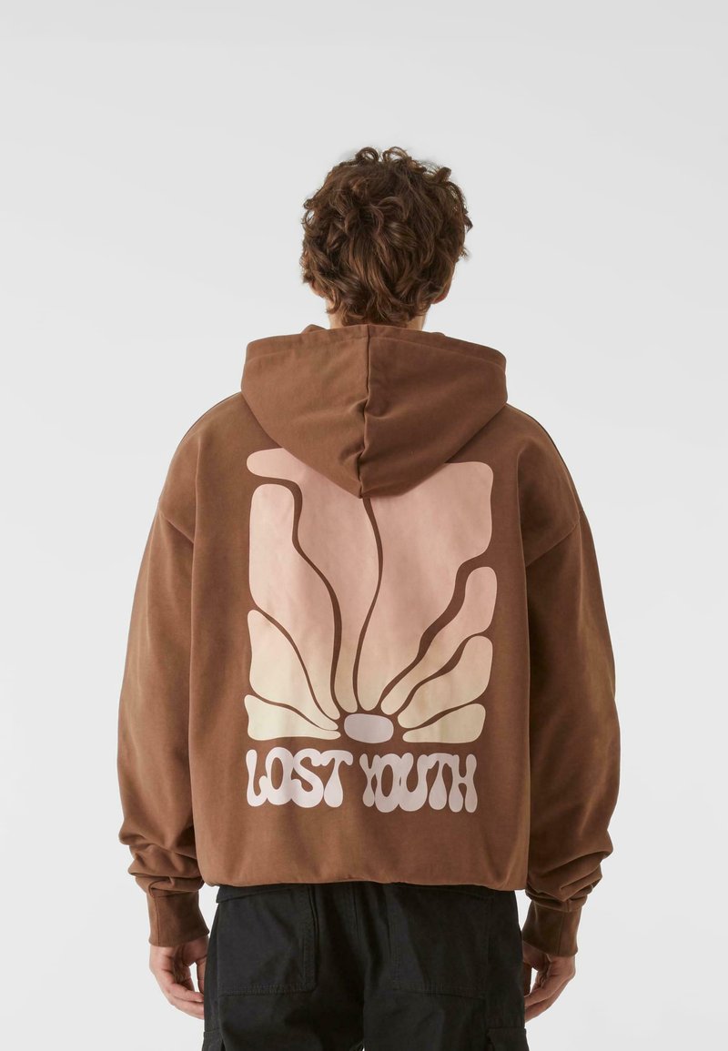 Barna kapucnis pulóver, amelyen egy nagy, stilizált pillangó grafika látható rózsaszín és krém színekben, alatta a "LOST YOUTH" felirattal.