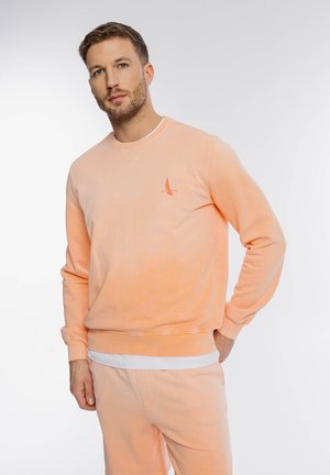 Mężczyzna z brodą w brzoskwiniowej bluzie i dopasowanych spodniach, stojący na białym tle z ręką w kieszeni.