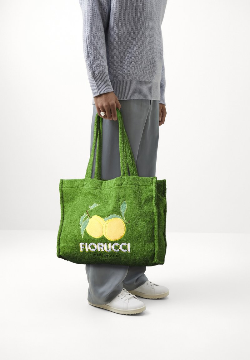 Personne portant un pull et un pantalon gris tient un sac fourre-tout FIORUCCI vert avec deux citrons jaunes et des feuilles, debout sur un fond blanc.