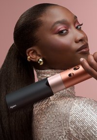 Dyson hårstylingsverktyg med svart kropp och rosaguldiga accenter, med en kamtillbehör och flera kontrollknappar, mot en rosa bakgrund.