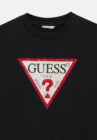 Svart bomull T-skjorte med et rødt og sølvtriangulært logo som har "GUESS® U.S.A." og et dekorativt spørsmålstegn i midten.