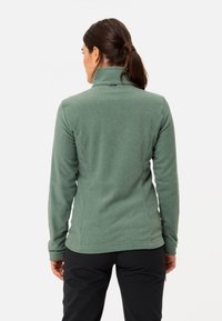 Giacca in pile verde con colletto alto, design slim fit e texture liscia. Caratteristiche includono un orlo arrotondato e maniche lunghe.