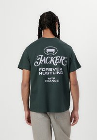 Tamsiai žalia medvilnė marškinėliai su laisvu siluetu, gale su dideliu baltu grafiniu tekstu, skaitant "JACKER FOREVER HUSTLING MTP FRANCE."