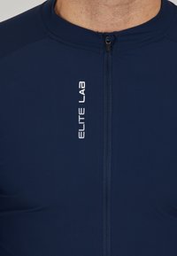 Navyblaues Sportoberteil mit Reißverschluss, das das Logo "ELITE LAB" in Weiß zeigt. Aus glattem, dehnbarem Stoff mit taillierter Passform gefertigt.