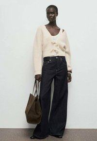 Cardigan a maglia crema con fiocchi decorativi, top bianco, pantaloni a gamba larga scuri e una borsa a spalla marrone, abbinati a scarpe nere.