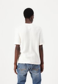 Weisses, gestricktes Kurzarmshirt mit vertikaler Rippstruktur, rundem Ausschnitt und lockerer Passform. Kombiniert mit blauen Jeans mit dekorativen Nähten.